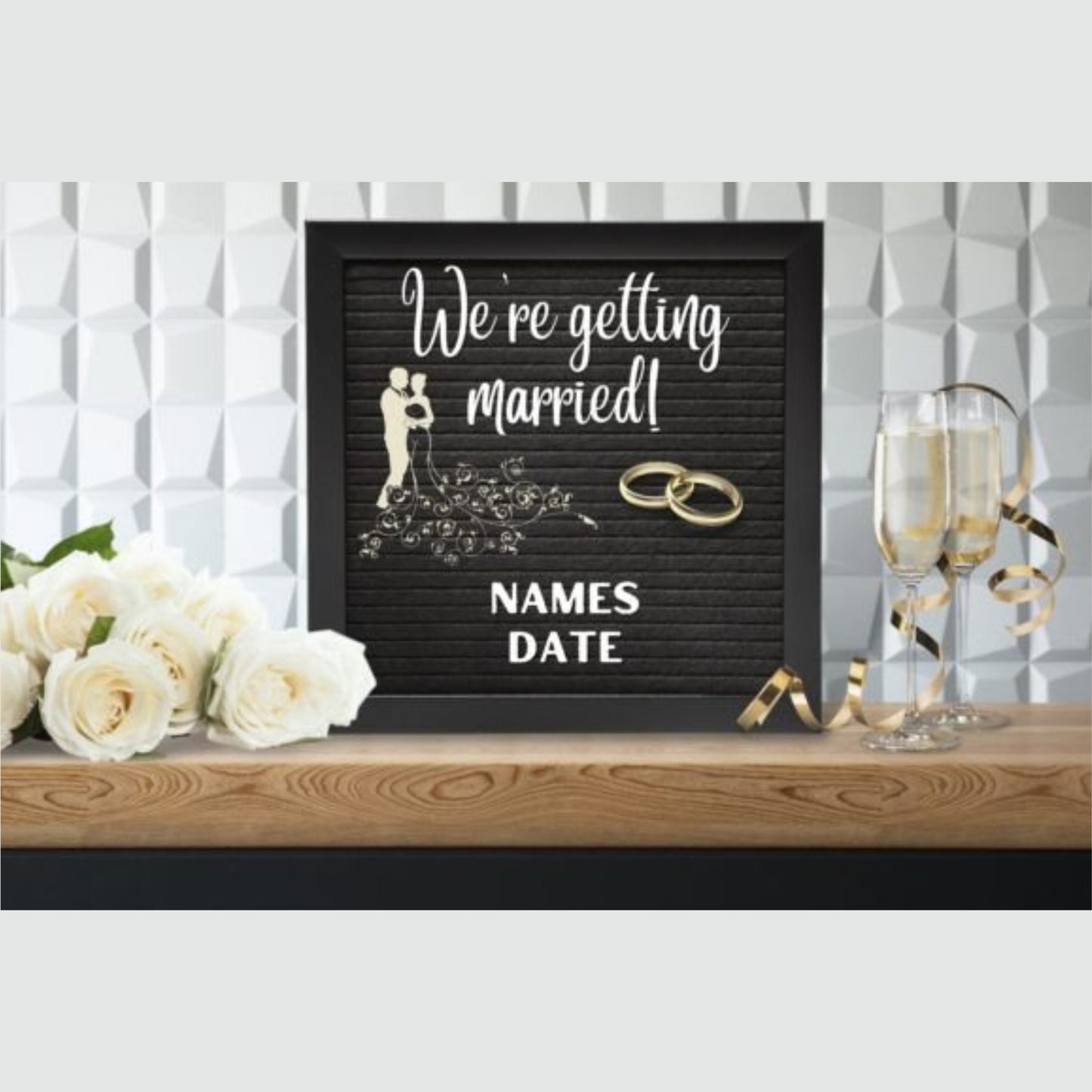 Black letter board with 'We're getting married!' text, wedding rings, and flowers on a wooden surface.