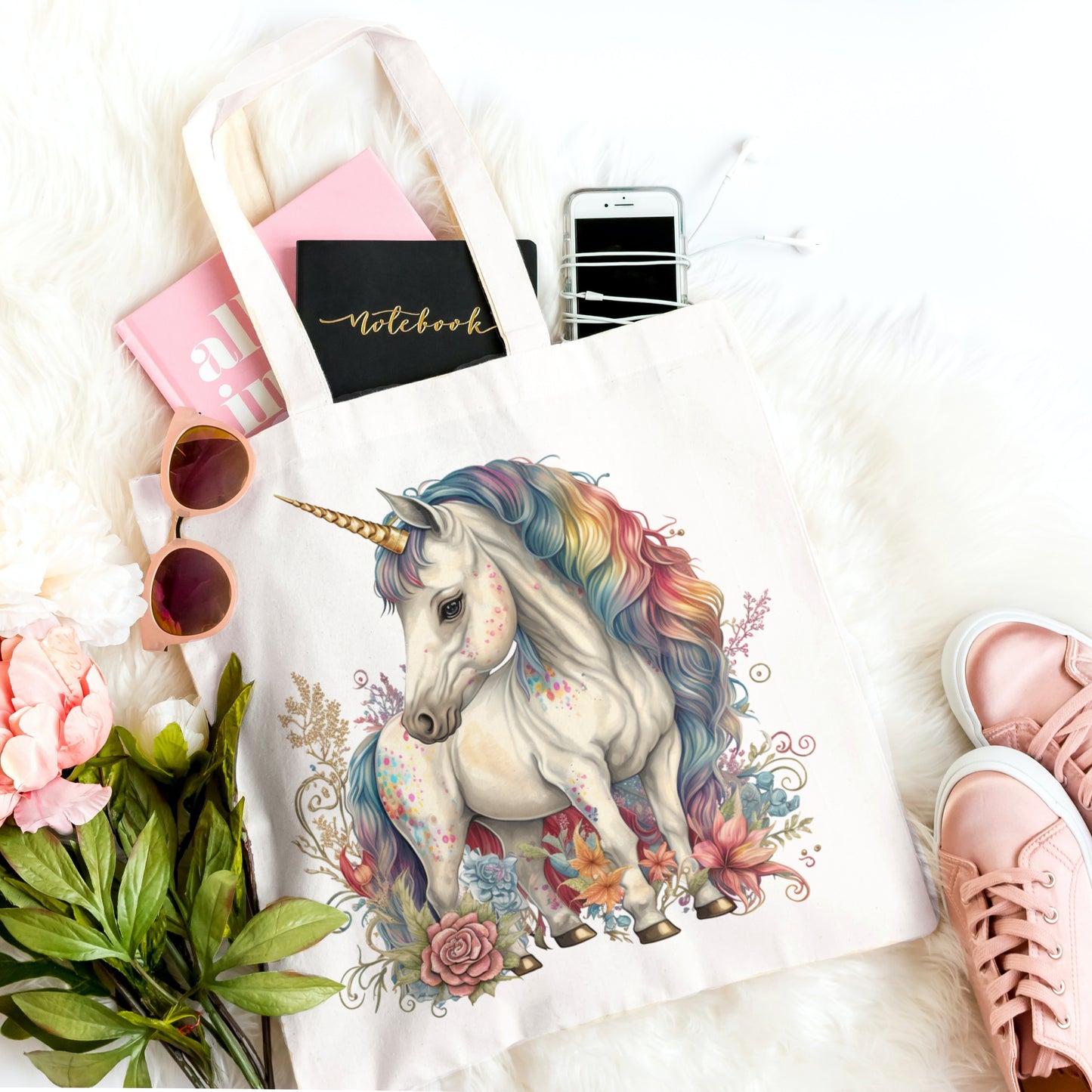 White tote bag with a colorful unicorn design, surrounded by phone, notebook, sunglasses, and flowers.