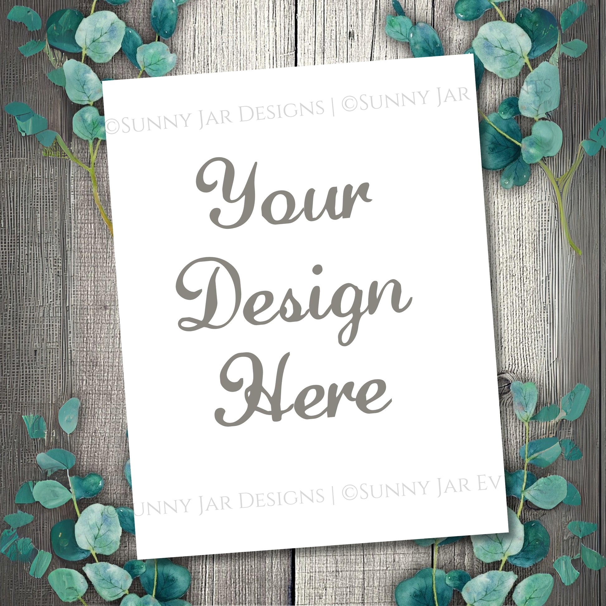 White paper with 'Your Design Here' text on a wooden background with eucalyptus leaves.