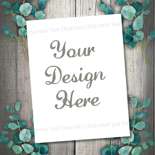 White paper with 'Your Design Here' text on a wooden background with eucalyptus leaves.