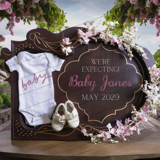 Decorative sign with baby onesie and shoes, 'We're Expecting Baby Jones May 2029' text, surrounded by flowers.