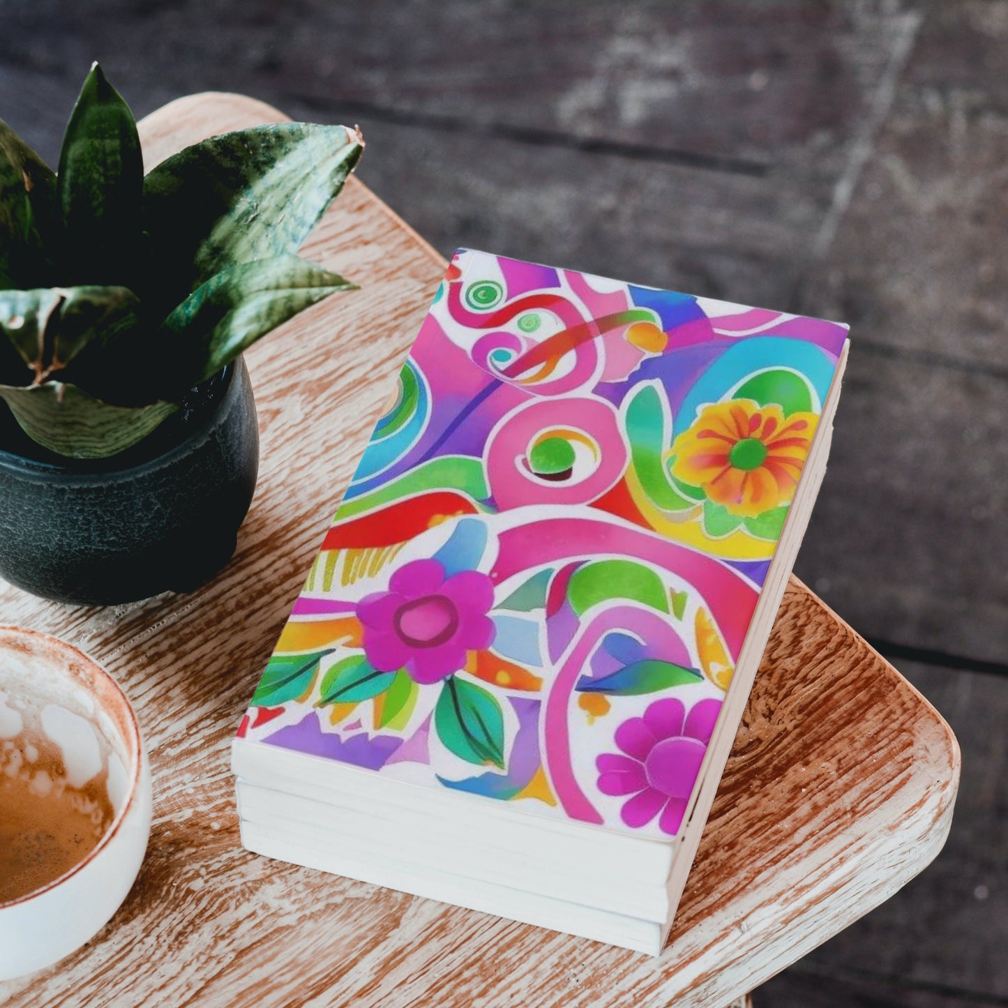Colorful notebook with floral design on a wooden table with a plant and cup of coffee.