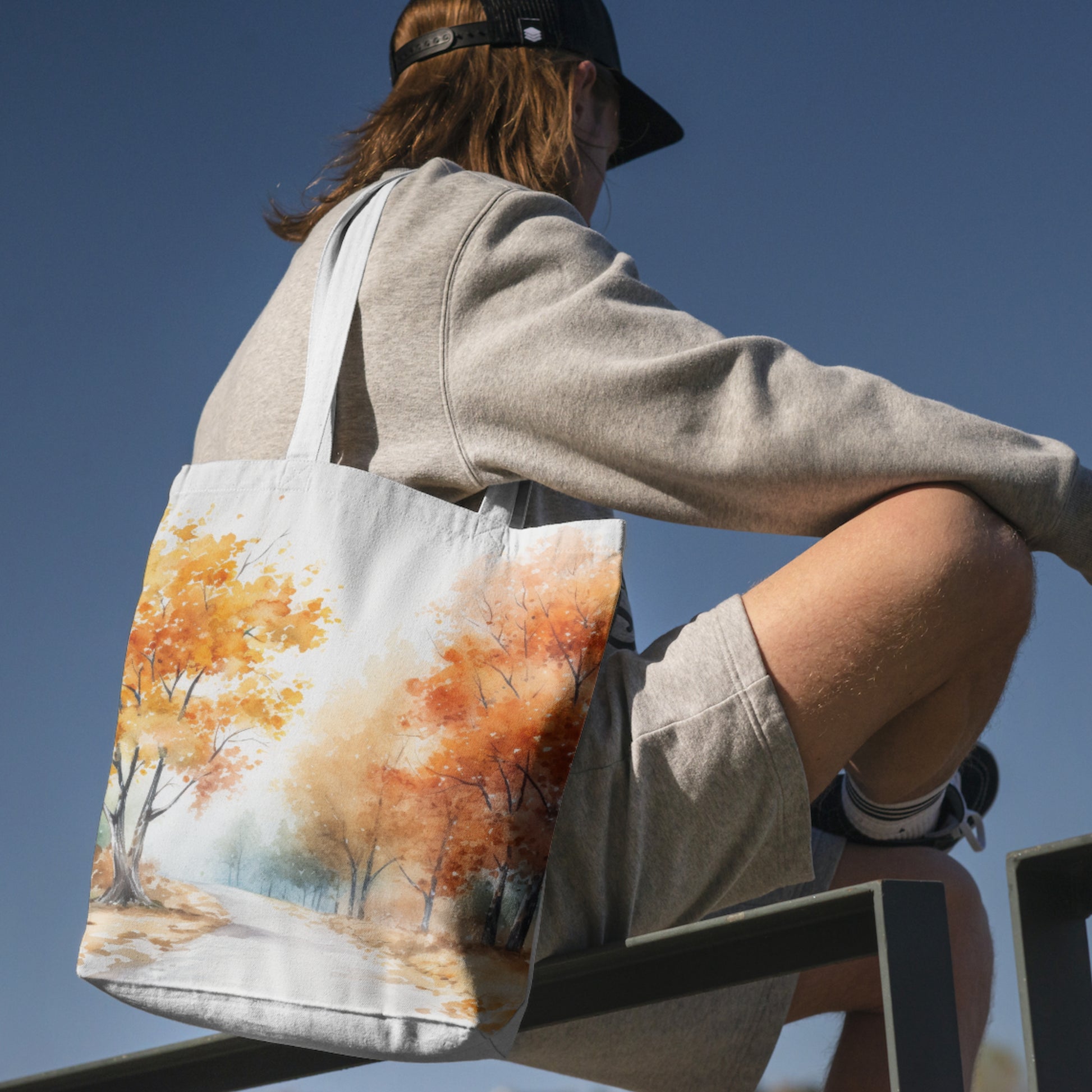 Person sitting with a tote bag featuring an autumn landscape design against a clear blue sky.