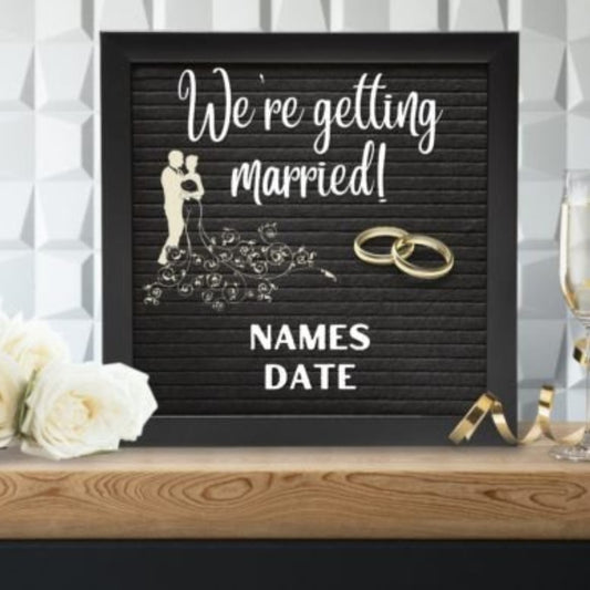 Black letter board with 'We're getting married!' text and wedding rings, placed on a wooden surface with flowers and glasses in the background.